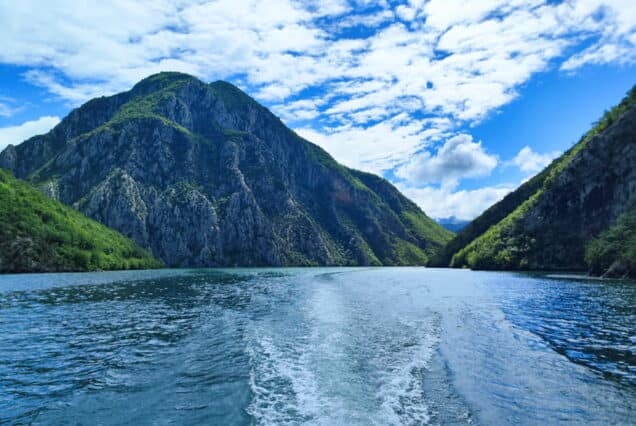 Boat wake on Komani Lake with mountain peak and blue sky