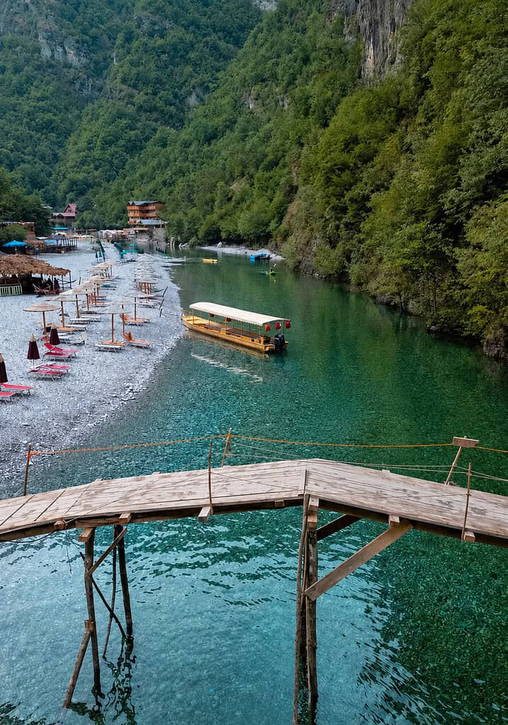 Wooden footbridge over the green water of Shala River with a tour boat below, Albania