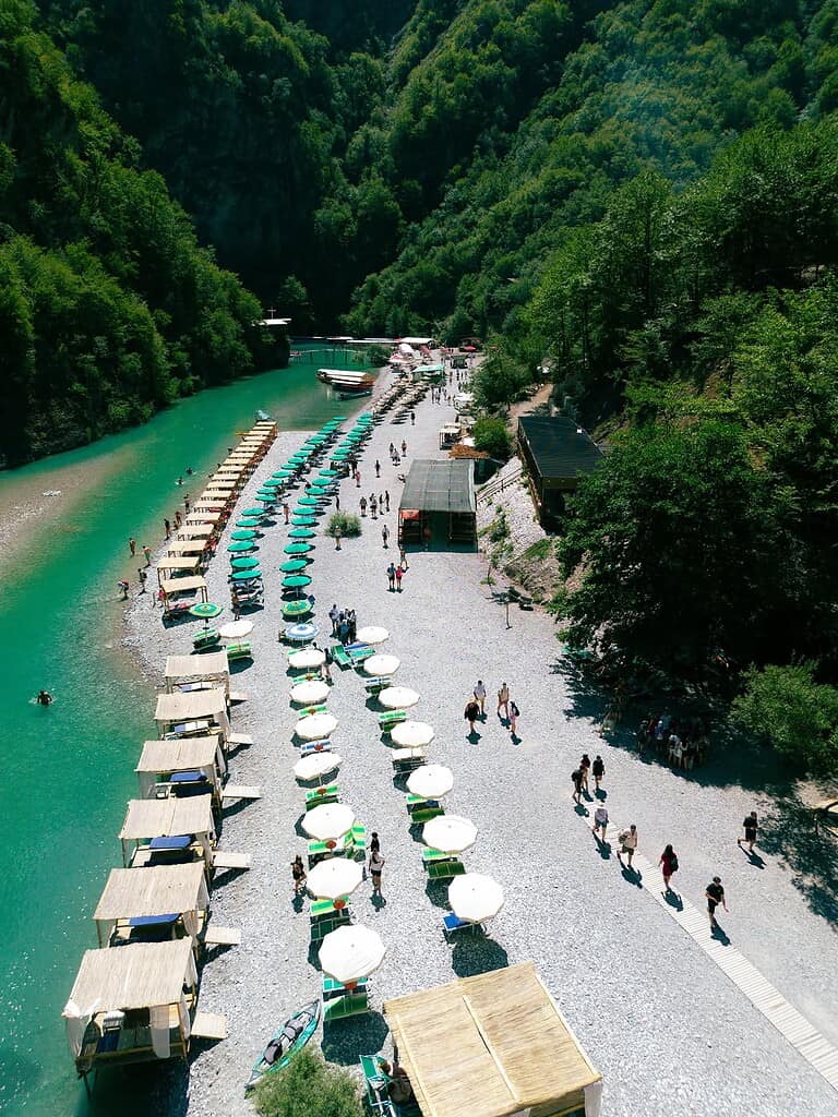 Aerial view of Shala River beach with turquoise water, sunbeds and mountain forest