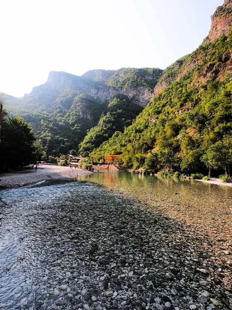 Clear shallow Shala River over pebbles in September, with green canyon walls and wooden cabins in the distance