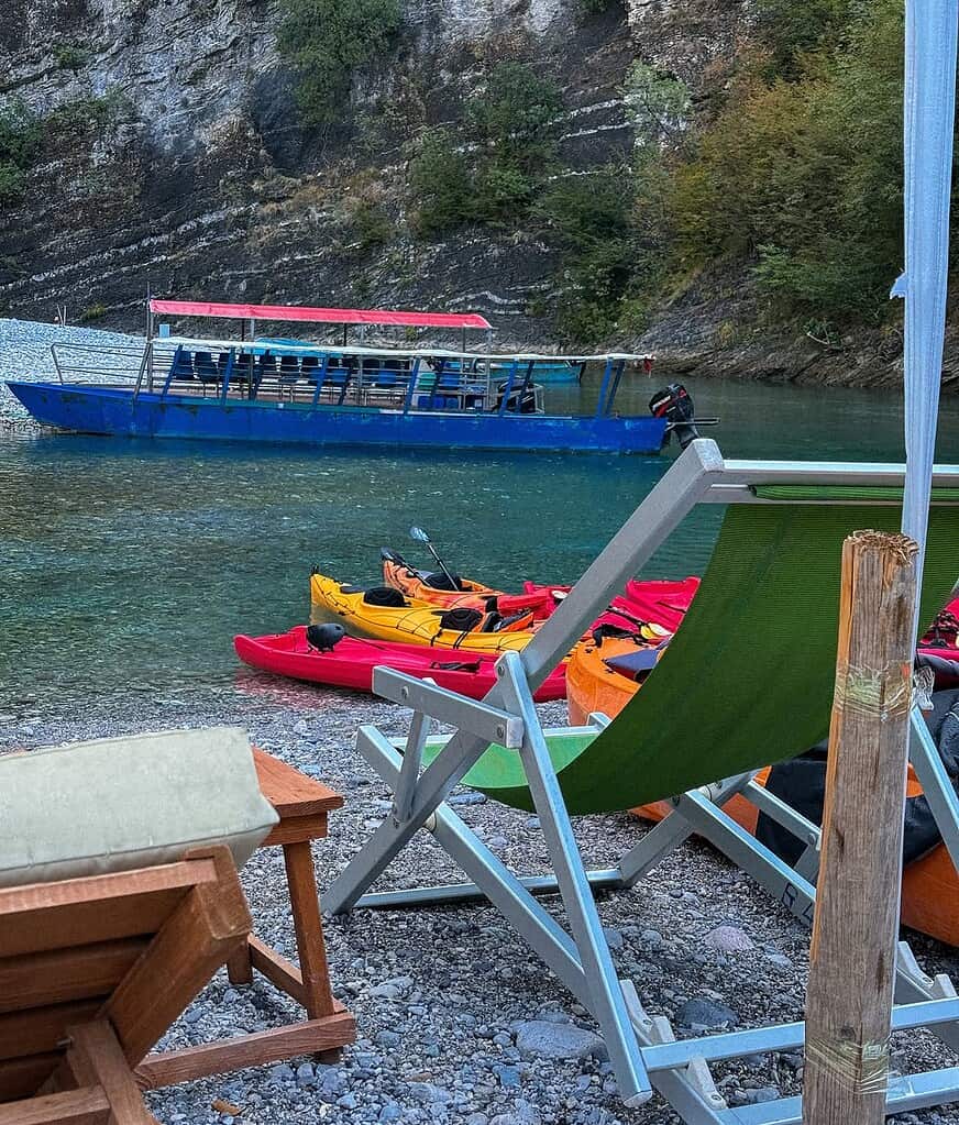 Colorful kayaks and a blue tour boat on the pebble shore of Shala River with a deck chair in front