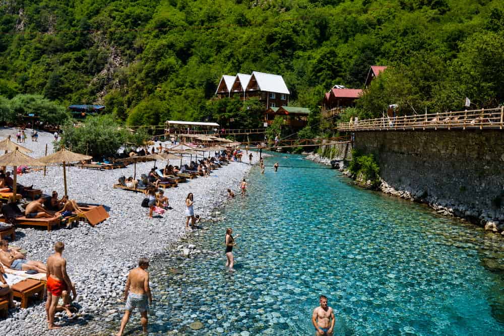 Visitors swimming near wooden guesthouses and a footbridge on Shala River, Albania