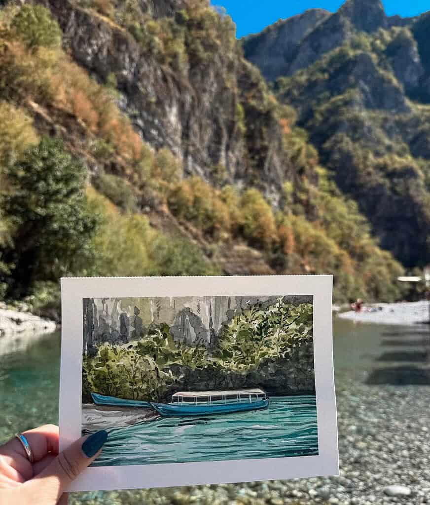 Hand holding a watercolor painting of boats on turquoise water in front of the real Shala River scene