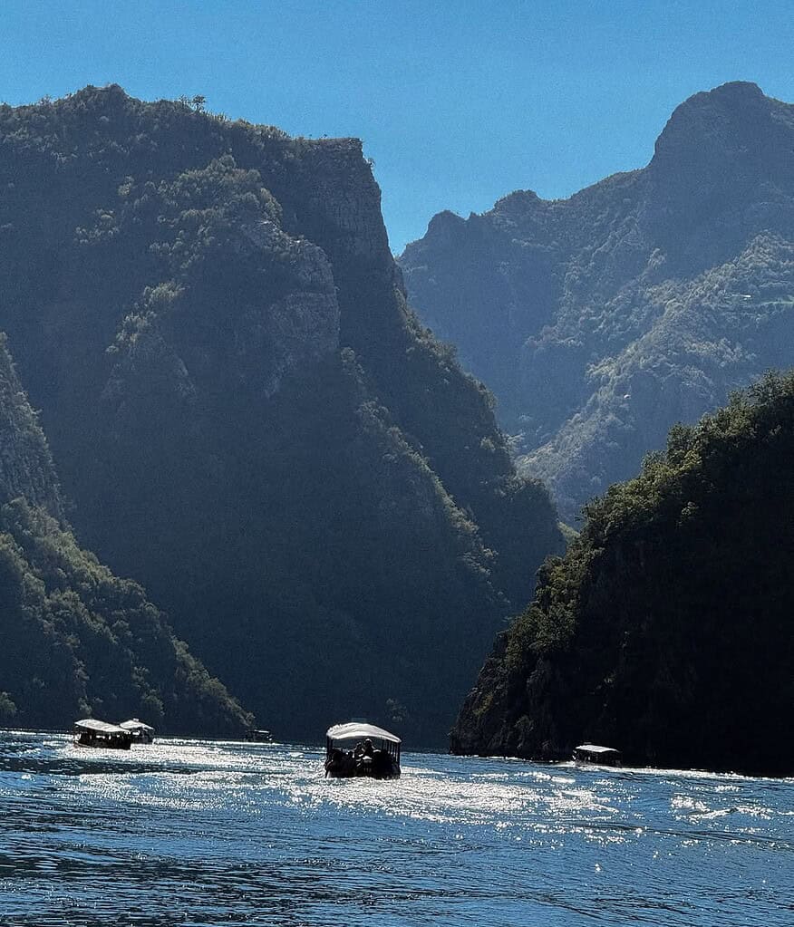 Tour boats crossing sparkling water between tall dark mountains on Koman Lake in September