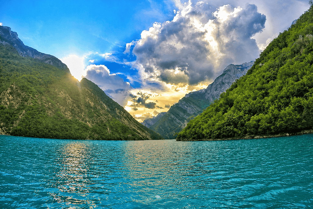 Turquoise Komani Lake with forested mountainsides and sun breaking through the clouds