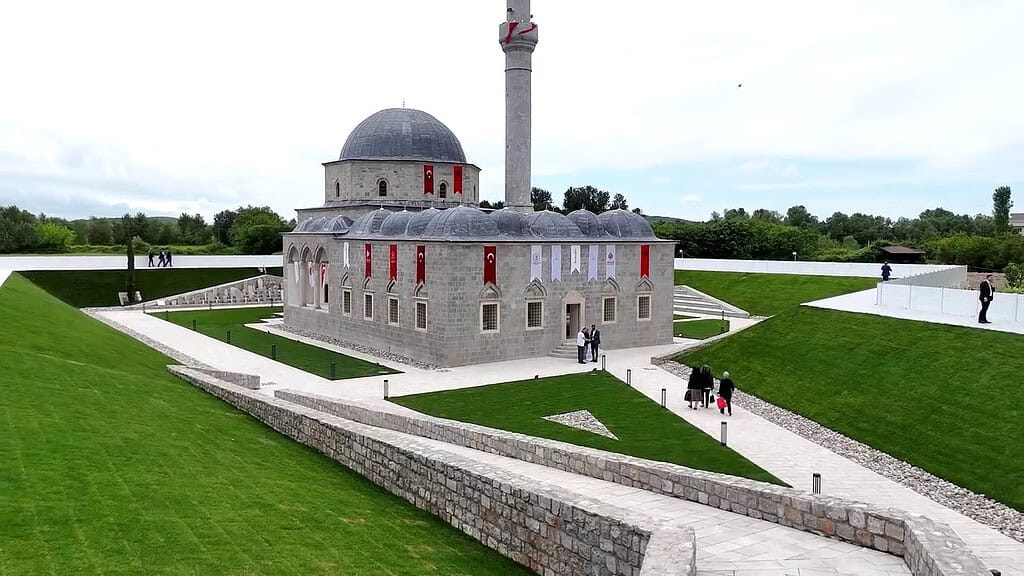 Ottoman-style stone mosque with gray domes and a tall minaret on landscaped grounds in Shkoder