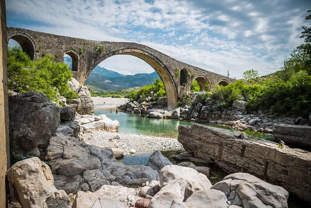 Long Ottoman stone arch bridge over turquoise river water with mountains visible through the arch
