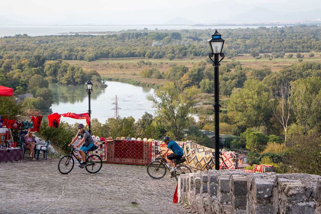 Two cyclists passing souvenir stalls with rugs and Albanian flags on a cobbled path above the river plain