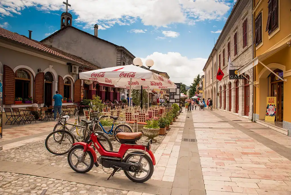 Red moped and bicycles parked beside café tables on a paved pedestrian street in Shkoder old town