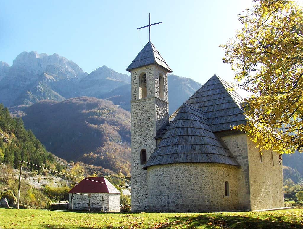 Stone church with a wooden-shingle roof and bell tower in Theth village below autumn mountains